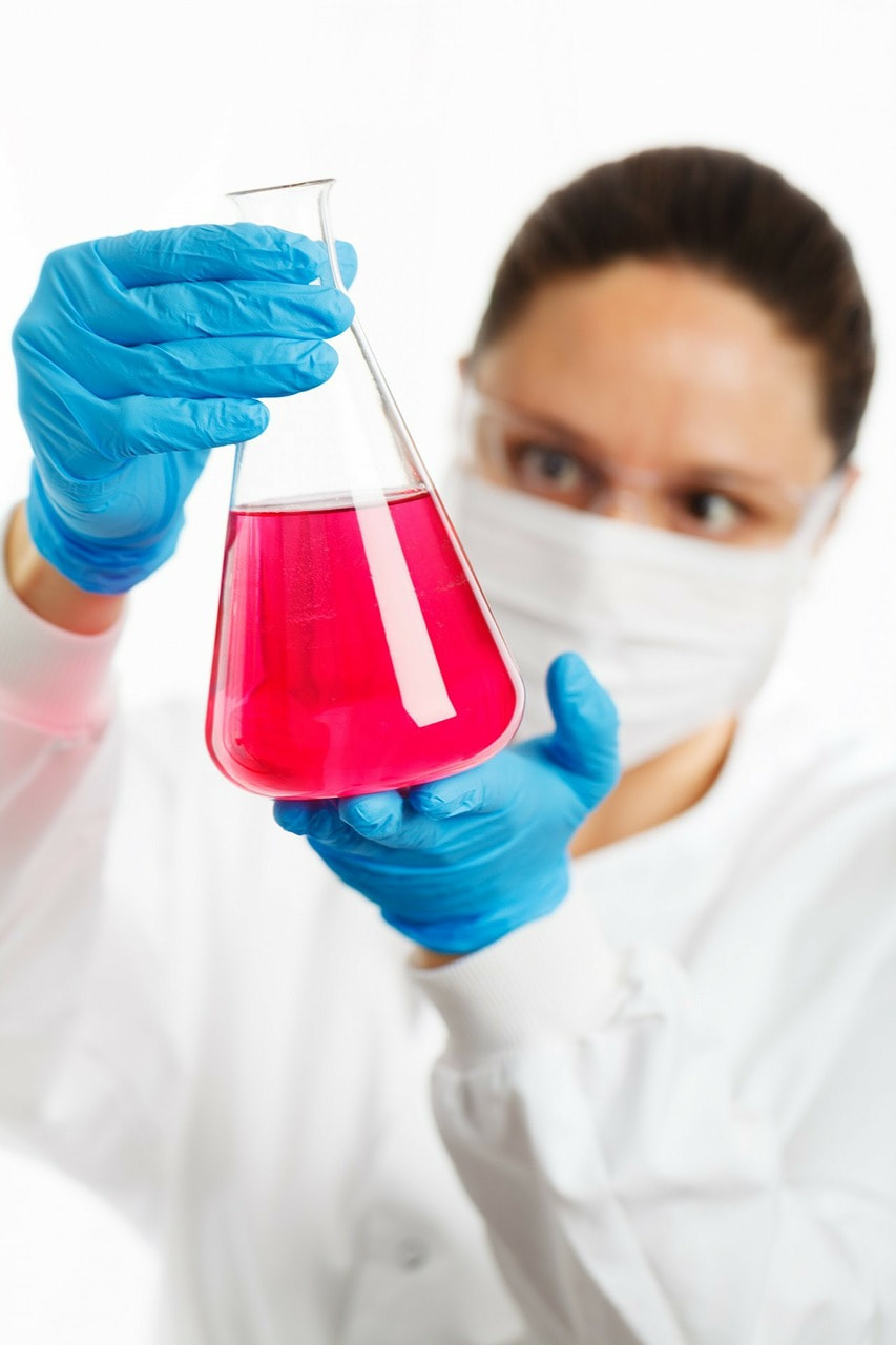 Woman in a lab coat looking at pink solution in an erlenmyer flask.
